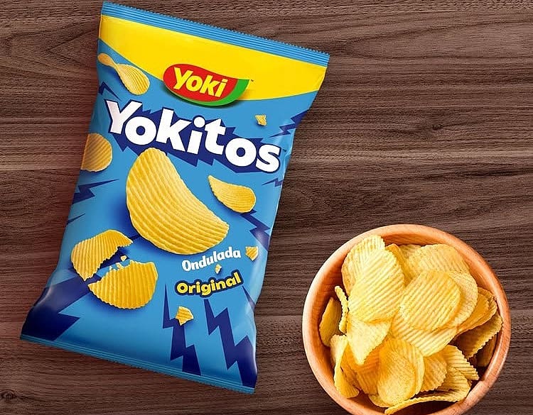 A bag of Yoki's Yokitos Ondulada Original chips next to a wooden bowl filled with ripple-cut chips on a wooden table.