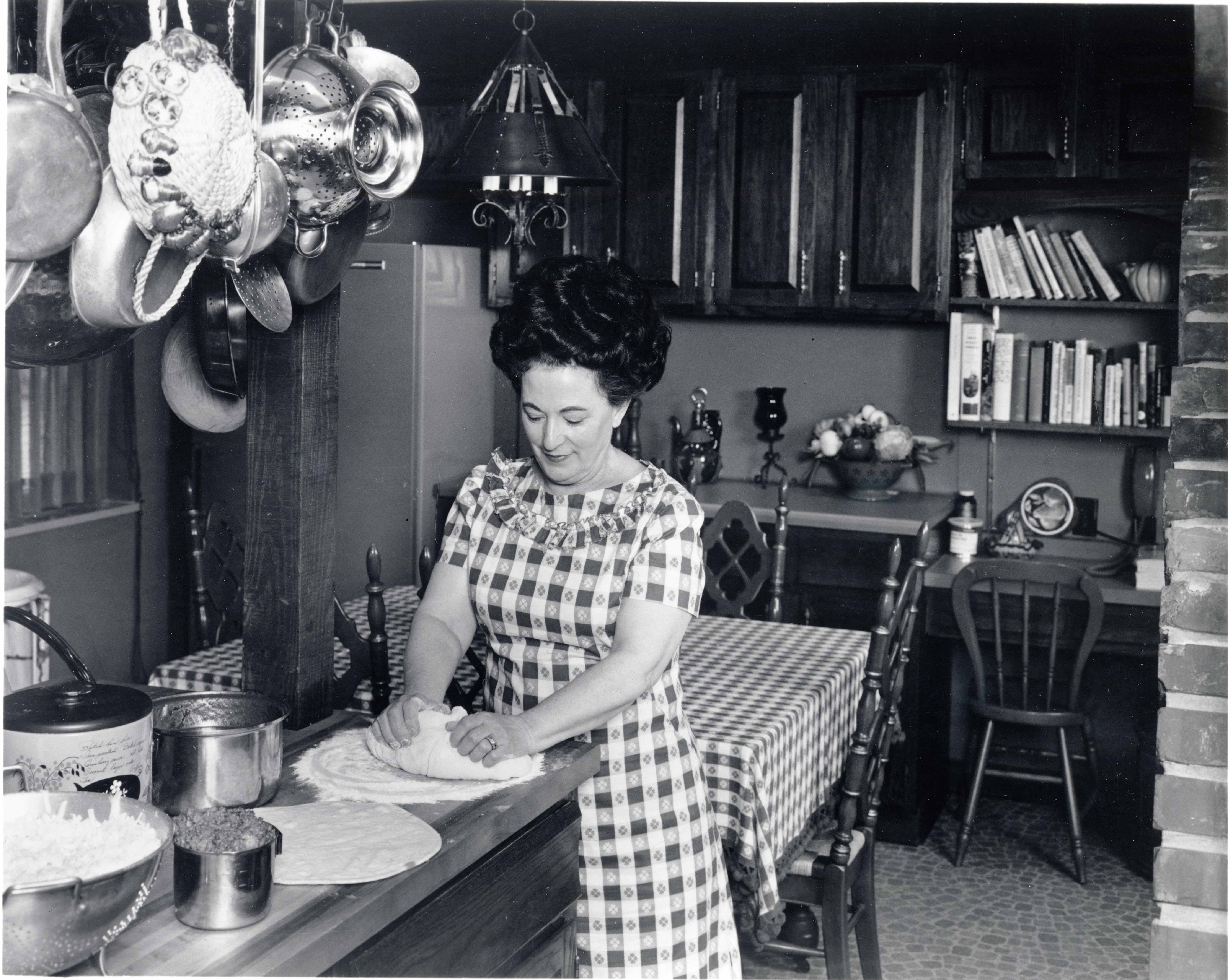 Black and white photo of Rose Totino wearing a checkered dress while hand kneading homemade pizza dough on her kitchen counter.
