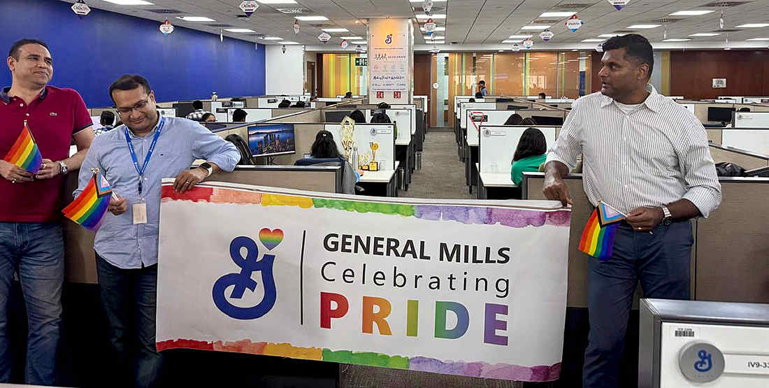 Mini Pride Walk inside the office- A group of people in cubicles holding a banner that reads "General Mills Celebrating Pride" with the General Mills logo displayed.