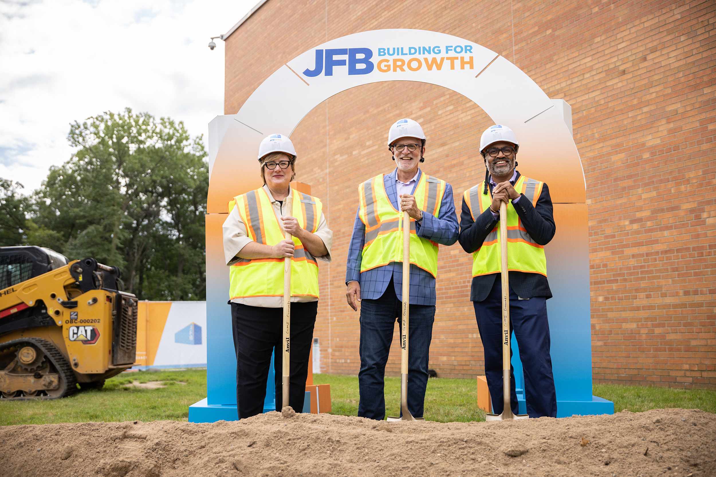 Three people standing in front of an arch that says "JFB: Building for Growth" wearing reflective vests and hard hats.