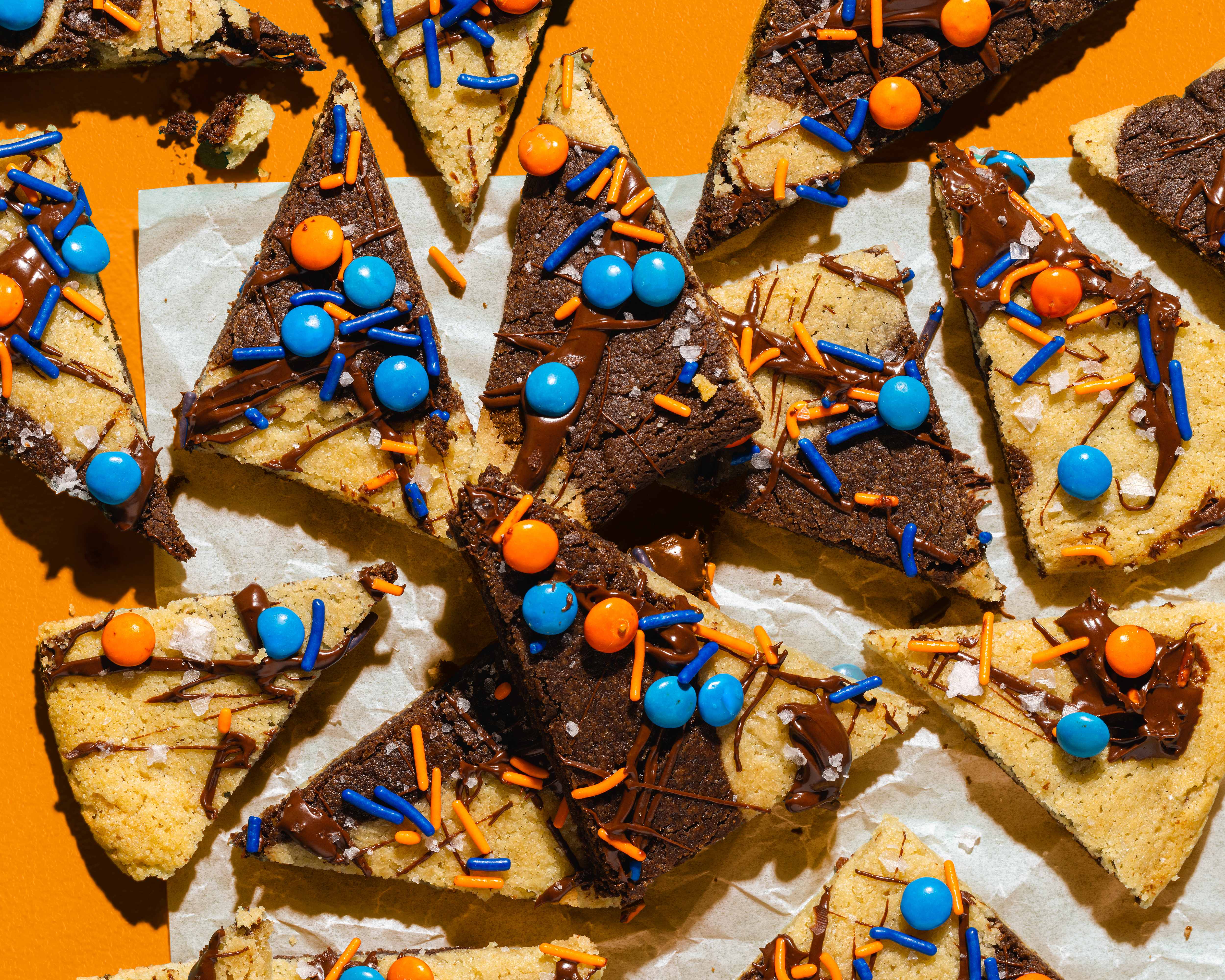 o	An overhead, close-up shot of triangular-shaped "Big Win Cookie Bark" dessert scattered on a piece of parchment paper against a solid orange background. The bars are made with marbled sugar cookie dough and cocoa powder, drizzled with melted chocolate and topped with blue and orange sprinkles, round candies, and a sprinkle of sea salt.