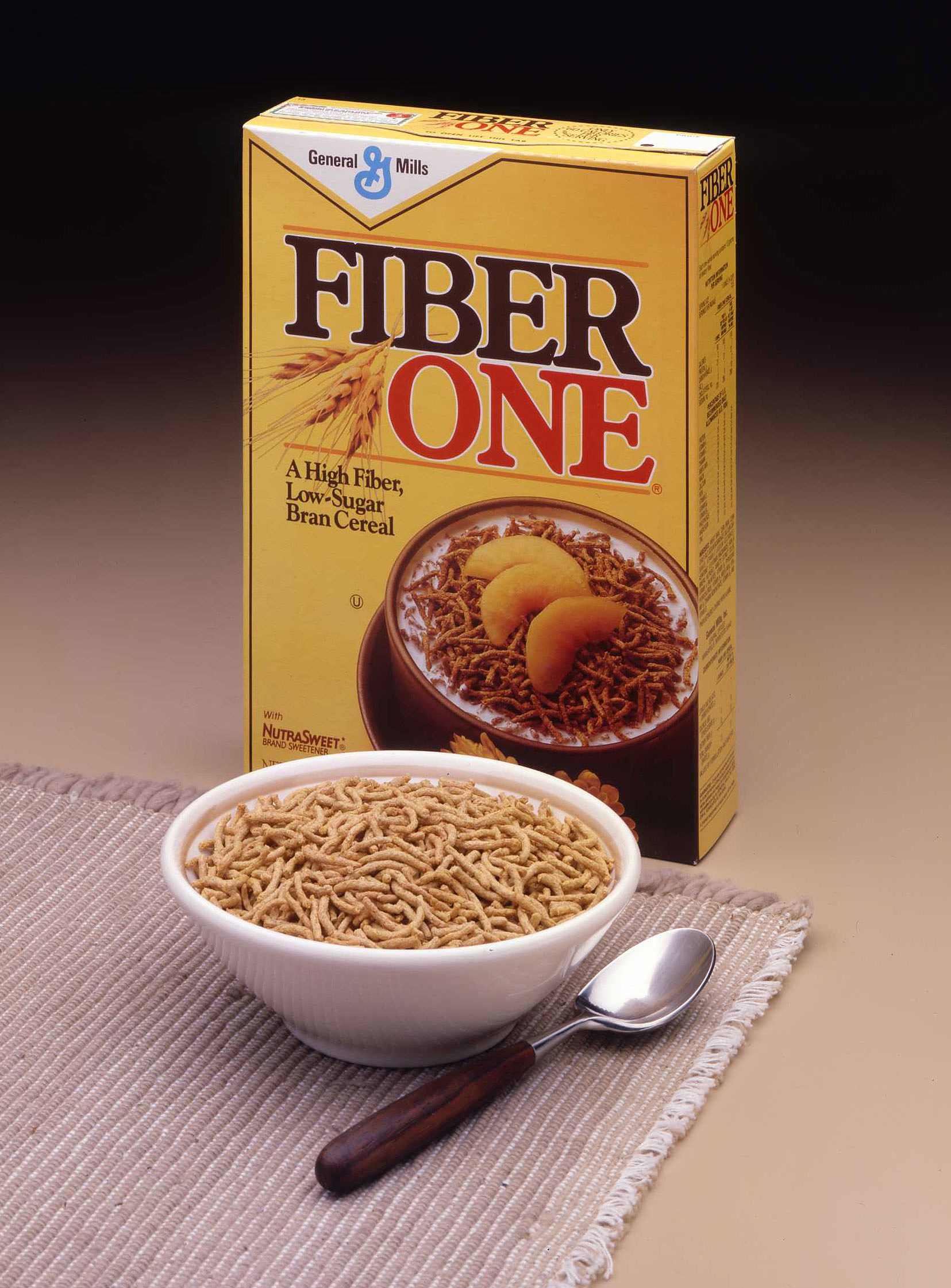 A classic product shot showing a yellow box of original Fiber One bran cereal. In front of the box, there is a full white bowl of the cereal placed on a textured placemat, with a spoon resting next to it.