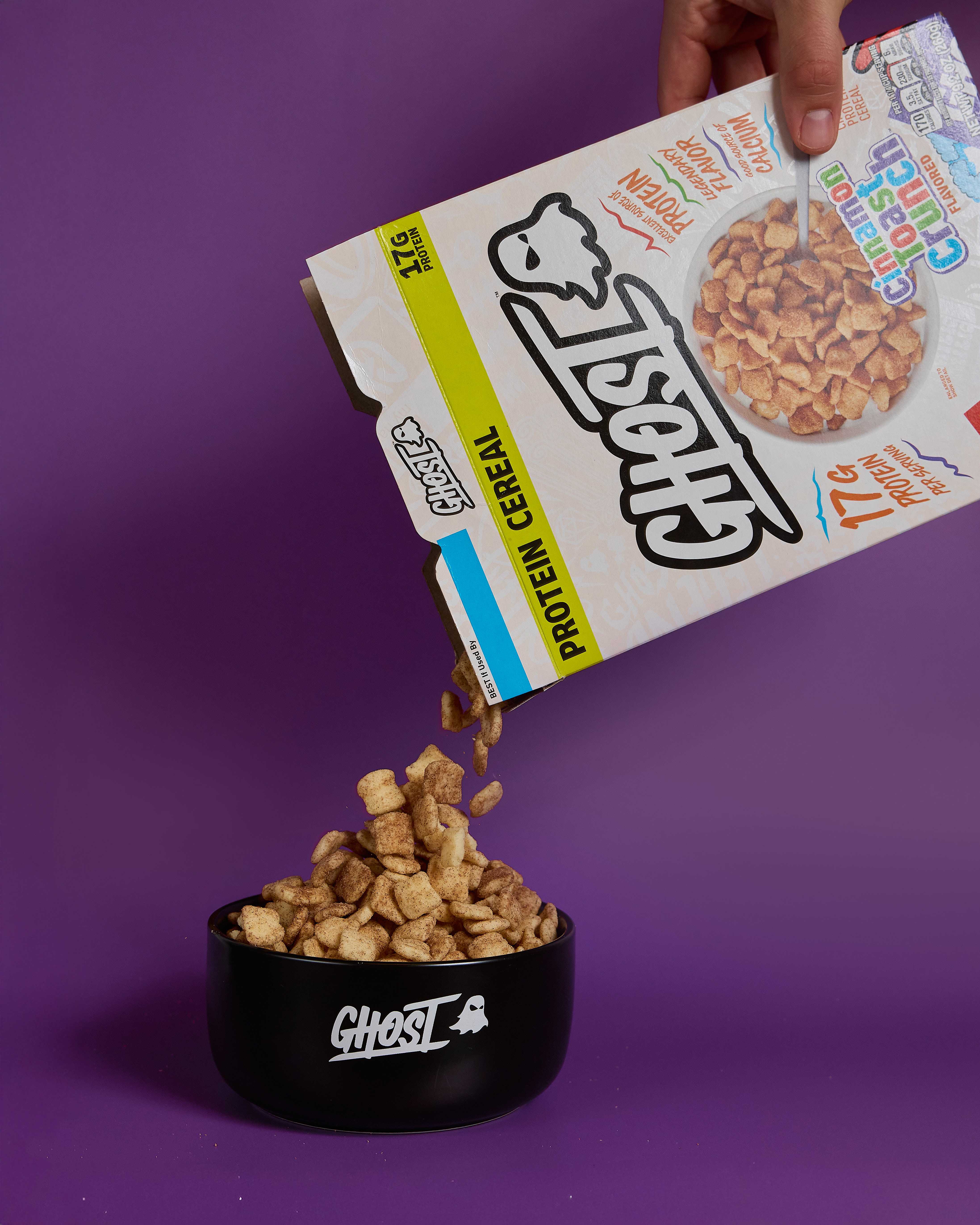 GHOST® PROTEIN CEREAL x CINNAMON TOAST CRUNCH™ being poured into a black bowl with a purple background.