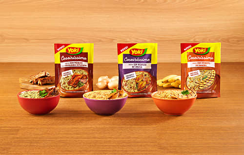 Three Yoki Brand Caseríssima seasoning packs with bowls of seasoned noodles, vegetables, and spices in front, against a wooden backdrop.