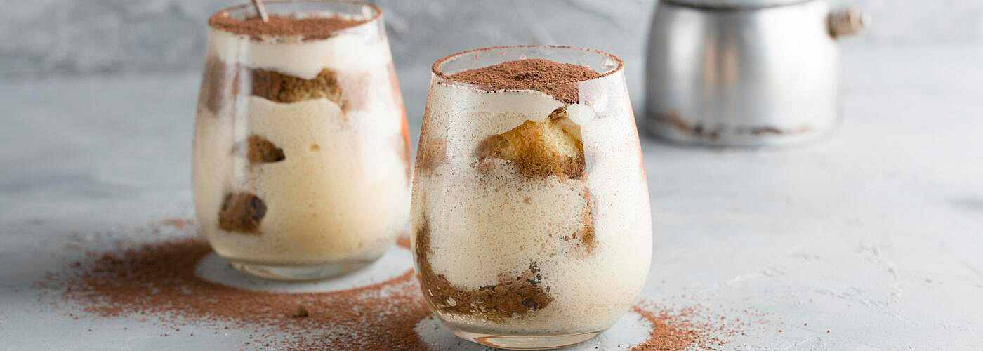 Two glasses of creamy tiramisu layered with mascarpone and soaked cake pieces, topped with a dusting of cocoa powder, placed on a light gray surface with cocoa scattered around.