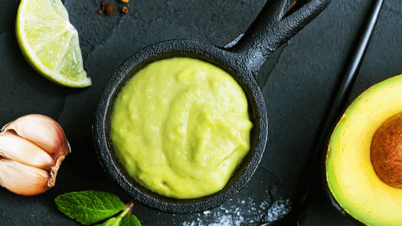 black bowl filled with creamy avocado sauce, surrounded by a lime wedge, halved avocado & garlic cloves on dark background