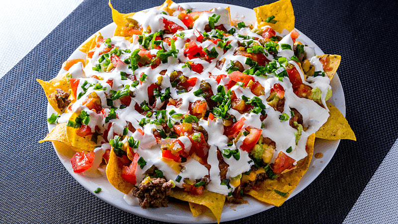 cheesy baked nachos topped with sour cream, diced tomatoes, coriander & other garnishes, served in a white plate on a black background