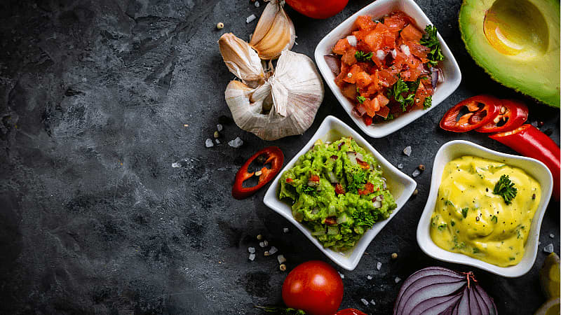 assorted mexican salsas including guacamole salsa, tomato salsa served on a rustic background with garlic, red chili, halved avocado around