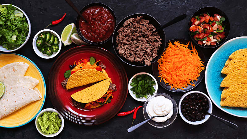 a taco bar with hard & soft shells, seasoned meat, cheese, salsa, beans, guacamole, sour cream & fresh toppings