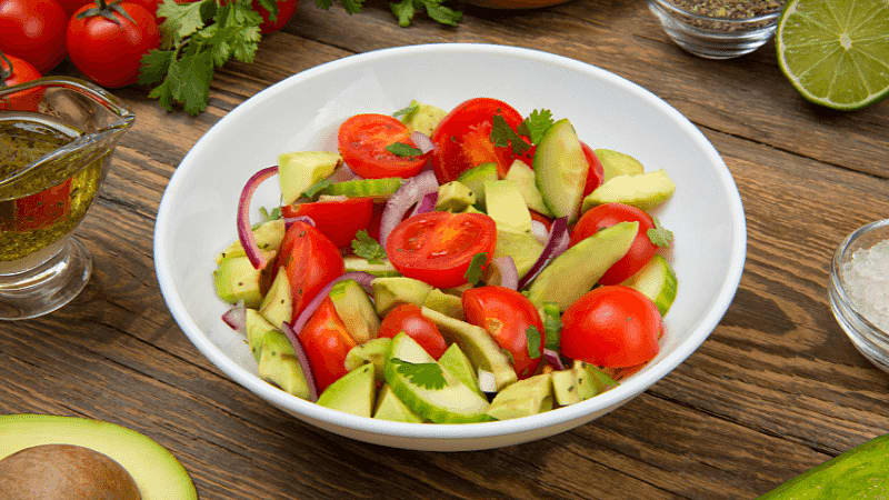 a white bowl filled with a fresh tomato avocado salad, garnished with sliced red onions & herbs, placed on rustic wooden table with ingredients in the background