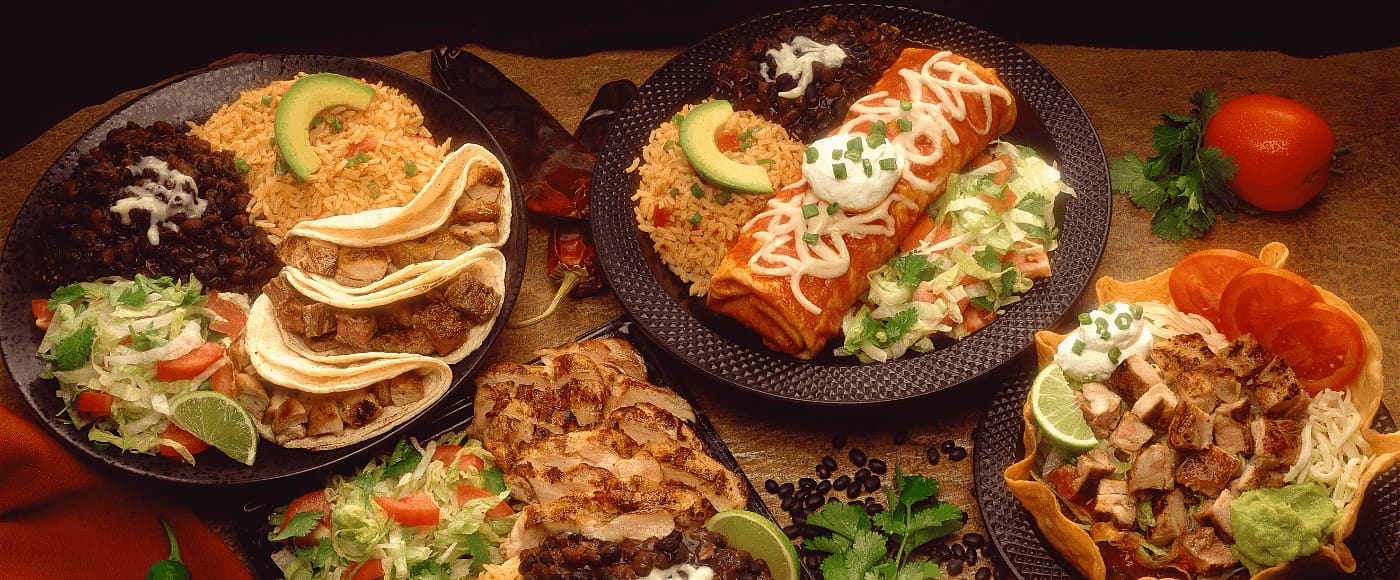 Mexican dishes with tacos, enchiladas, rice, beans & salad, garnished with avocado, lime, sour cream & fresh veggies