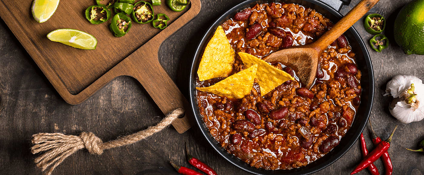 skillet filled with chili beans, minced meat & tortilla chips surrounded by jalapenos, lime wedges and chili peppers on a rustic wooden board