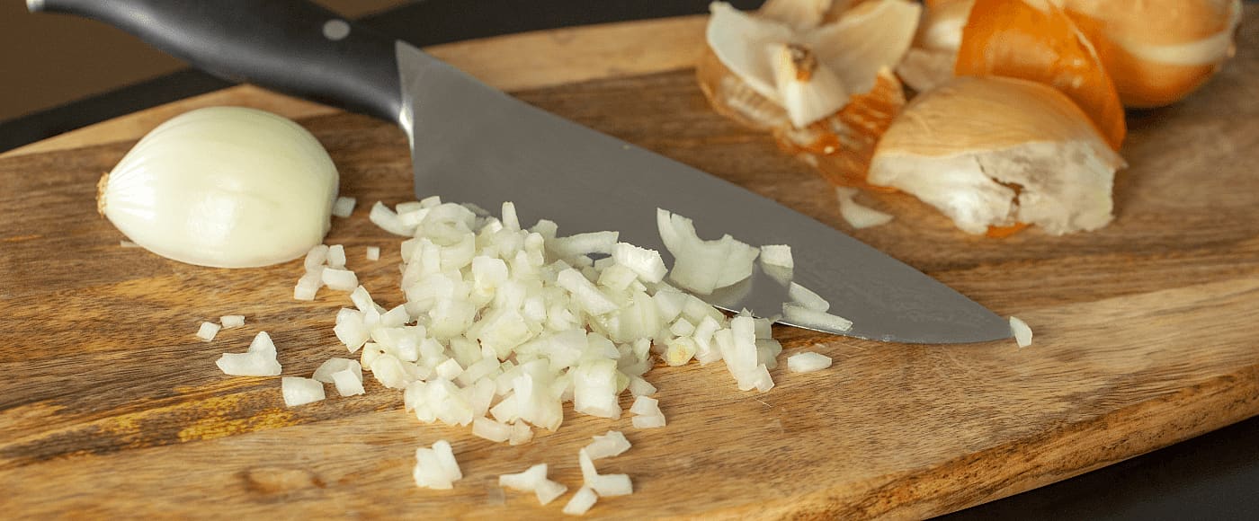 chopped onions on a wooden cutting board with kitchen knife, partially peeled onion & onions skins in the background