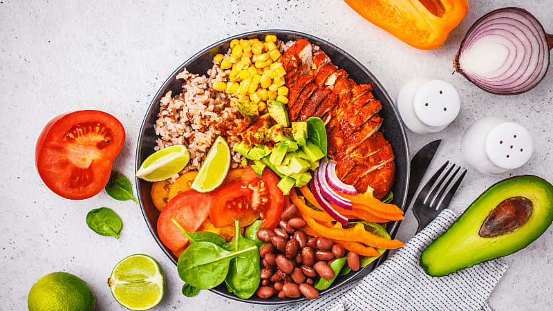 a vibrant bowl of ingredients such as chicken, beans, avocado, corn & fresh veggies styled with lime & seasonings