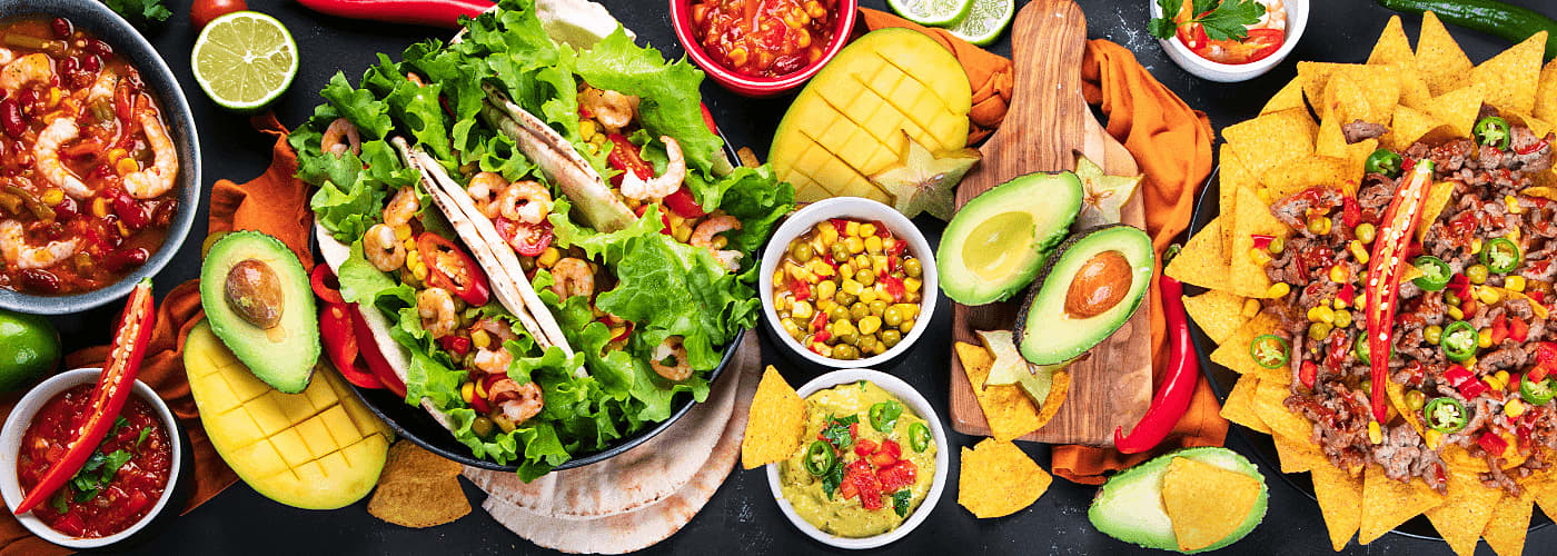 an assortment of vibrant Mexican inspired dishes including tacos, machos, guacamole, salsa, avocado, mango and decorative garnishes on a dark background