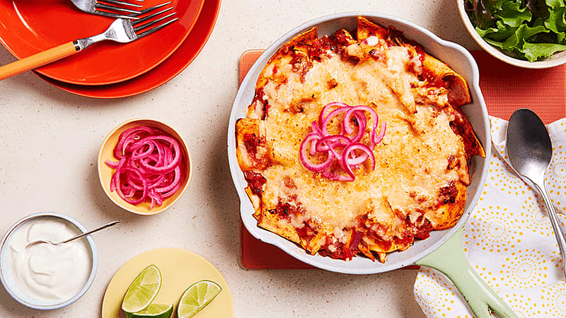 One Pan Chicken Enchiladas in a skillet surrounded with a bowl of greens, red onion, soured cream and lime wedges
