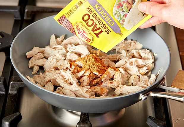 Shredded chicken in a skillet on the stove with a hand pouring a packet of Old El Paso Chicken Taco seasoning, front of package, over the chicken